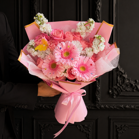 A bouquet of pink and white flowers wrapped in pink paper and tied with a pink ribbon, held by a person wearing a black suit against a dark ornate door.